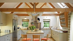 Una cocina amplia con una isla central, sillas de madera, un refrigerador de acero inoxidable y una ventana con vista a la vegetación.