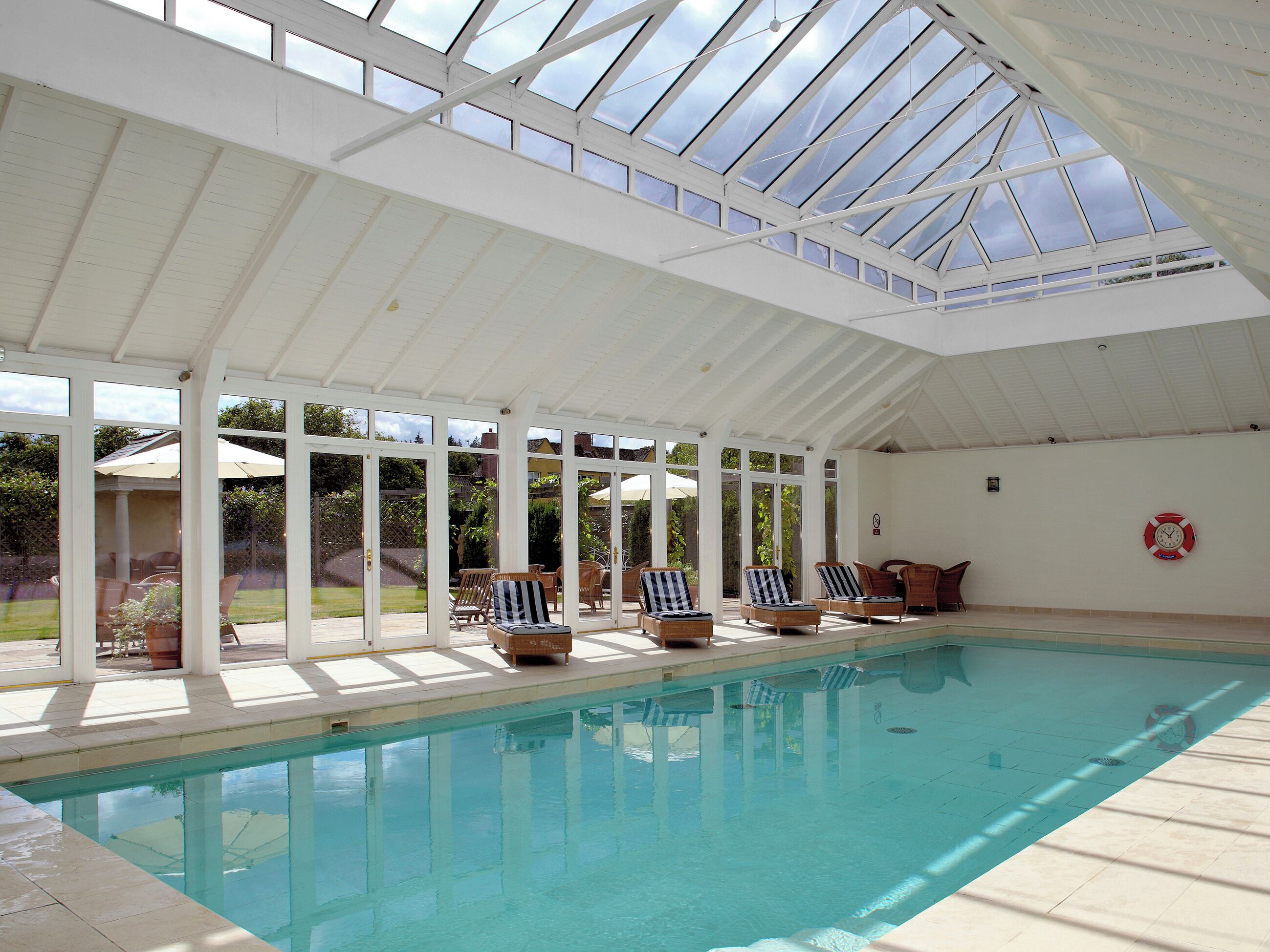 An indoor swimming pool with a glass ceiling, lounge chairs, and a lifebuoy on the wall.