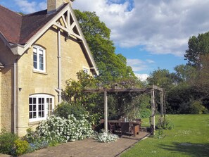 Ein Steinhaus mit Satteldach, einer Holzpergola und einem Garten mit blühenden Pflanzen.