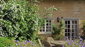A brick house with a window and a door, surrounded by lush greenery and flowering plants. - 3 bedroom accommodation in Bruern, near Chipping Norton (Bruern, near Chipping Norton)