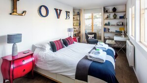 Een slaapkamer met een bed, een nachtkastje in rood, een boekenplank en een raam.