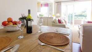 A dining table set with a bottle of wine, fruit, and glassware, with a view of a living area and outdoor space. - 1 bedroom accommodation in Thorpeness, near Aldeburgh (Thorpeness, near Aldeburgh)