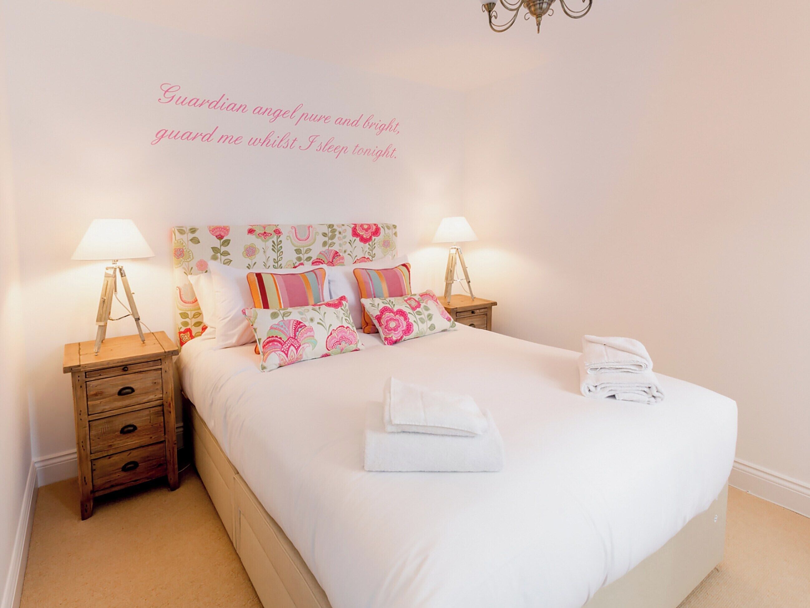 A neatly made bed with floral-patterned pillows and a white comforter, flanked by two bedside tables with lamps and a small wooden drawer.