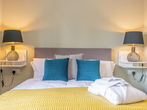 A neatly made bed with a yellow textured bedspread, two blue pillows, and a white blanket folded at the foot of the bed. There are two wall-mounted lamps with black shades on either side of the bed. - 5 bedroom accommodation in Dunholme, near Lincoln (Dunholme, near Lincoln)