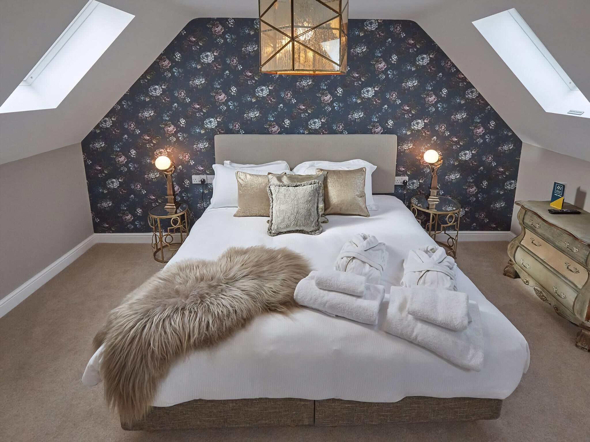 Een zolderkamer met een schuine kap, een groot bed met wit beddengoed, twee bedlampen en een bont plaid.