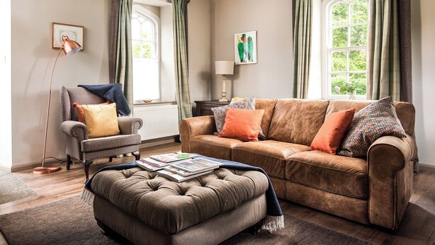 A cozy living room with a brown leather sofa, a patterned armchair, and a matching ottoman. There are two windows with green curtains, a floor lamp, and a table with a lamp and books. - 2 bedroom accommodation in Longframlington, near Rothbury (Longframlington, near Rothbury)