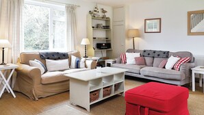 A well-lit living room with two sofas, a coffee table, and a red ottoman. There are two lamps, a bookshelf, and a framed picture on the wall.
