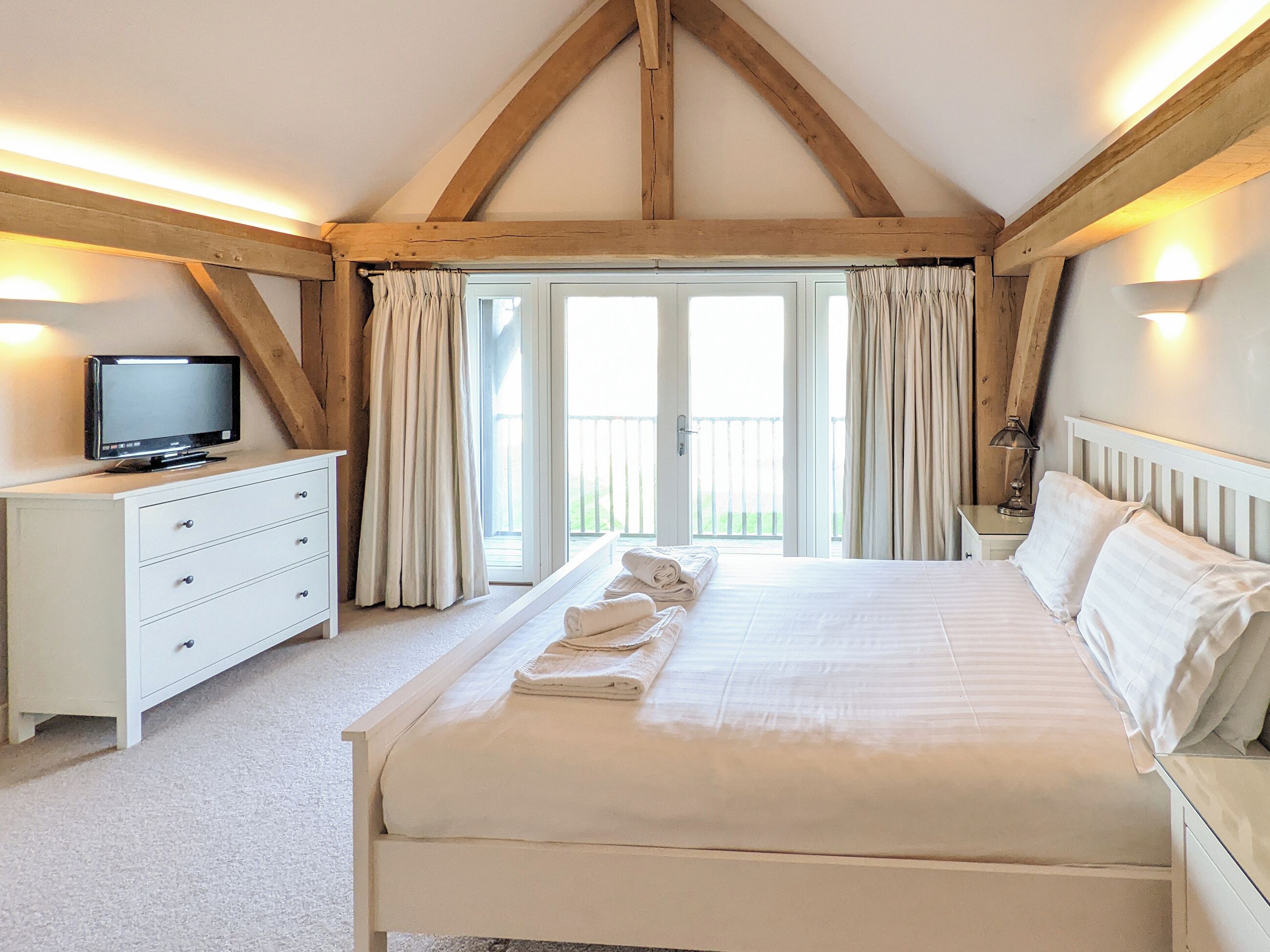 A bedroom with a large bed, a TV on a dresser, and a balcony with curtains.