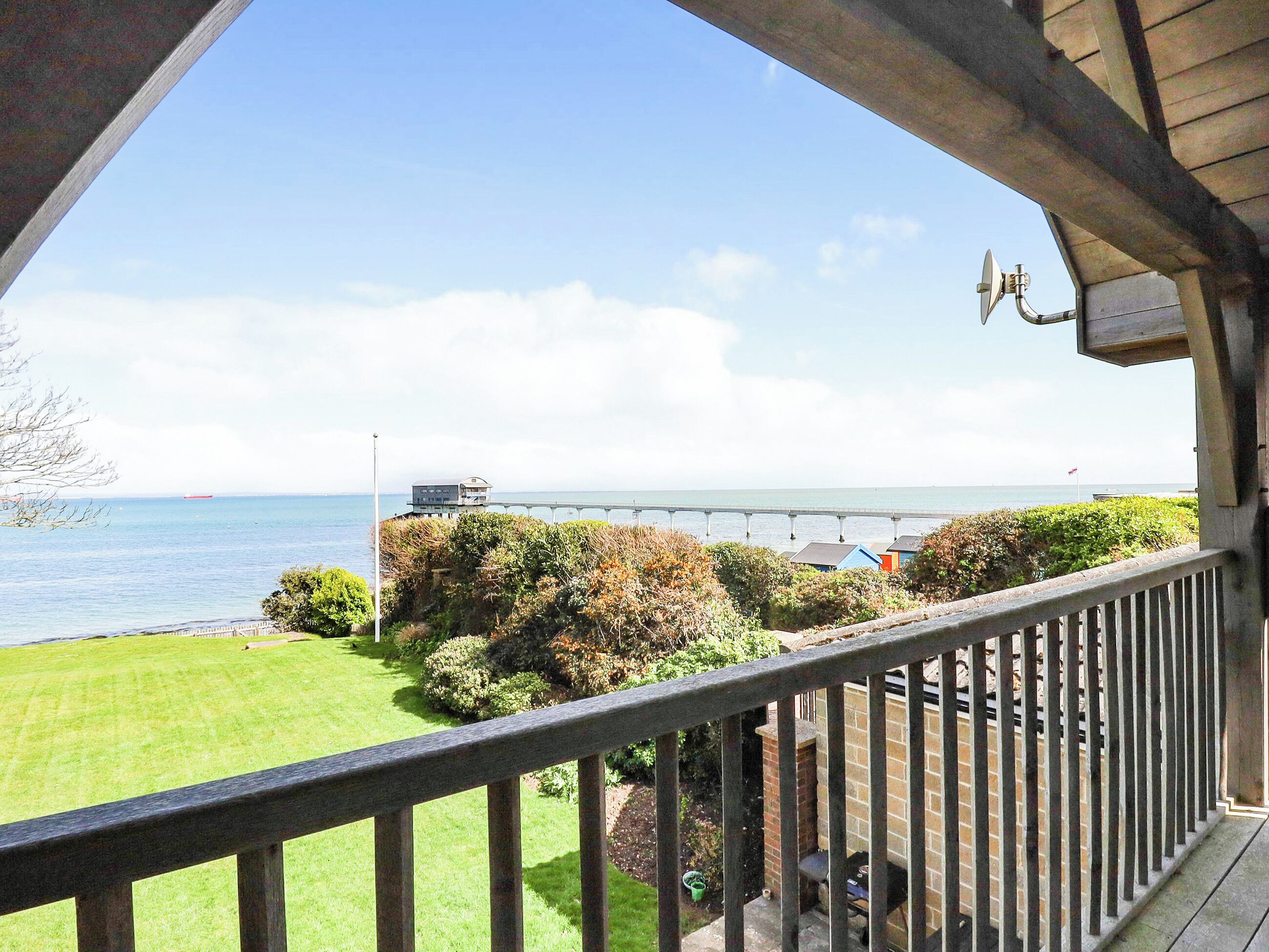 A balcony with a view of the sea, a pier, and a grassy area with bushes.