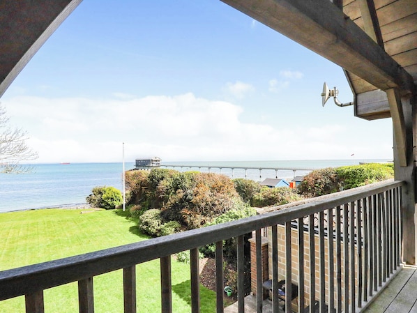 A balcony with a view of the sea, a pier, and a grassy area with bushes.