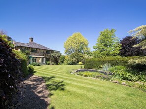 Une maison en pierre avec une pelouse bien entretenue, un patio couvert et une variété d’arbres et d’arbustes.