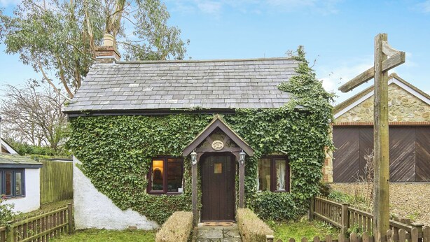 Une petite maison avec un chemin de pierre menant à une porte en bois, entourée d’une clôture en bois et de verdure.