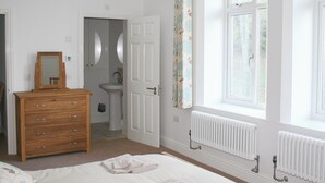 Une chambre à coucher comprenant un lit, une commode, un miroir, une fenêtre avec des rideaux, un radiateur et une porte donnant sur une salle de bain.