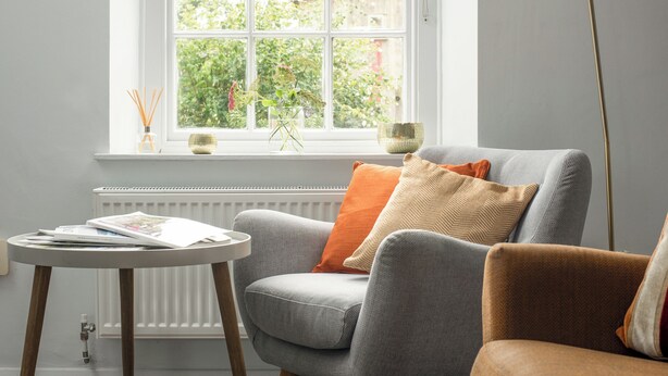 Een woonkamer met een grijze bank, een oranje kussen, een ronde tafel met een tijdschrift, een vloerlamp en een raam met uitzicht op groen.