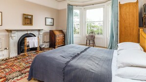 A bedroom with a large bed, a wooden dresser, a chair, a fireplace, and a window with curtains. - 6 bedroom accommodation in Brading, near Sandown (Brading, near Sandown)