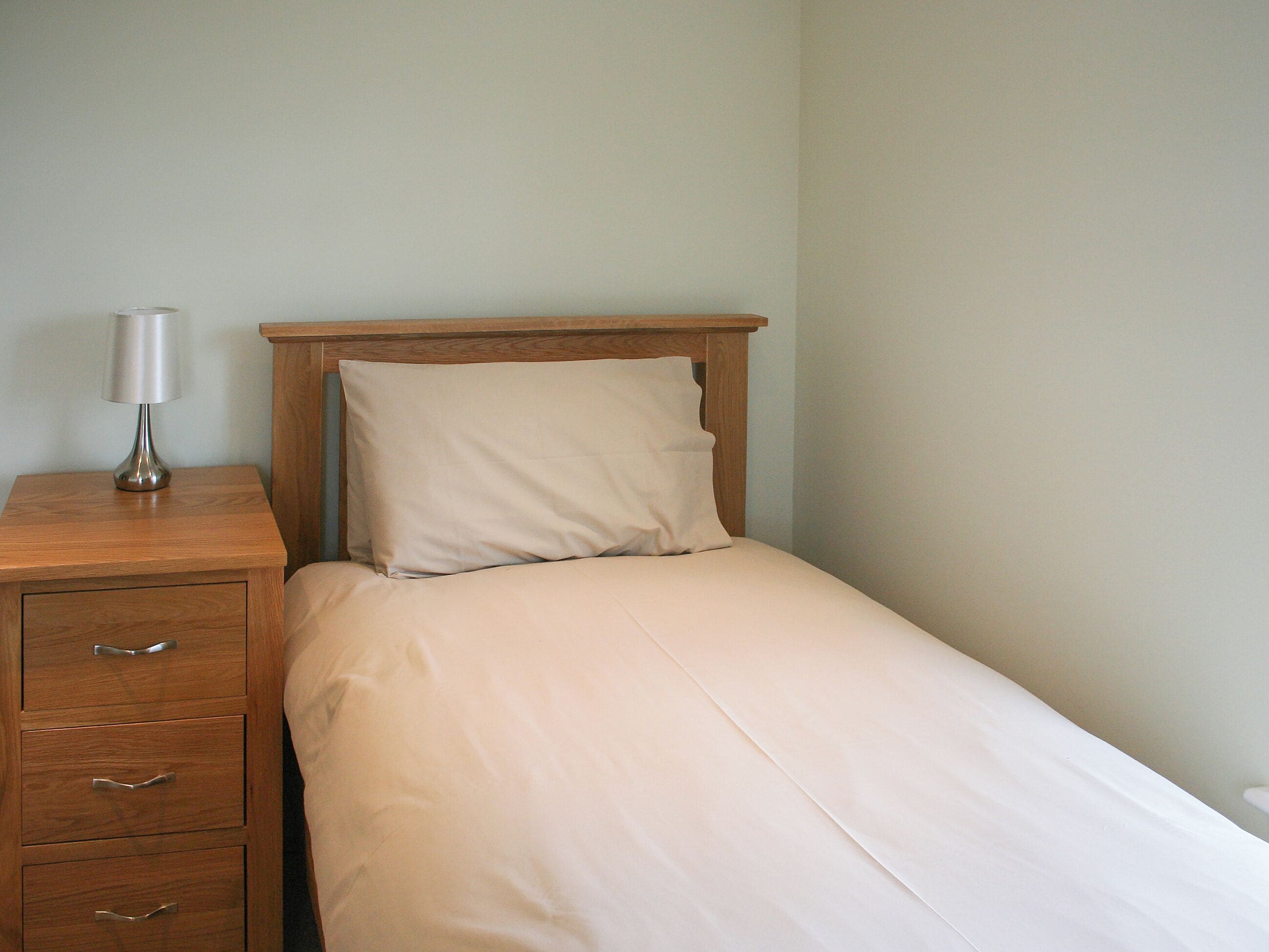 A small, neatly made bed with a wooden headboard, a bedside table with a lamp, and a drawer unit.