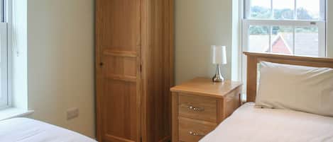 A bedroom with two beds, a wardrobe, a bedside table with a lamp, and a window with a view of a house.
