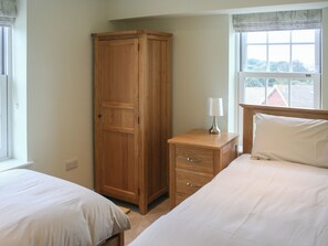 A bedroom with two beds, a wardrobe, a bedside table with a lamp, and a window with a view of a house. - Highlands Apartment 3 - UKC3105 (Shanklin)