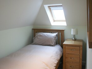 A bedroom with a sloped ceiling, a single bed with a wooden headboard, a bedside table with a lamp, and a small wooden dresser. - Highlands Apartment 4 - UKC3106 (Shanklin)