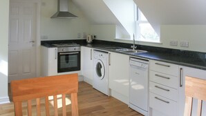 Cocina compacta con horno empotrado, lavadora y lavavajillas, una silla de madera y un ventanal que proporciona luz natural.