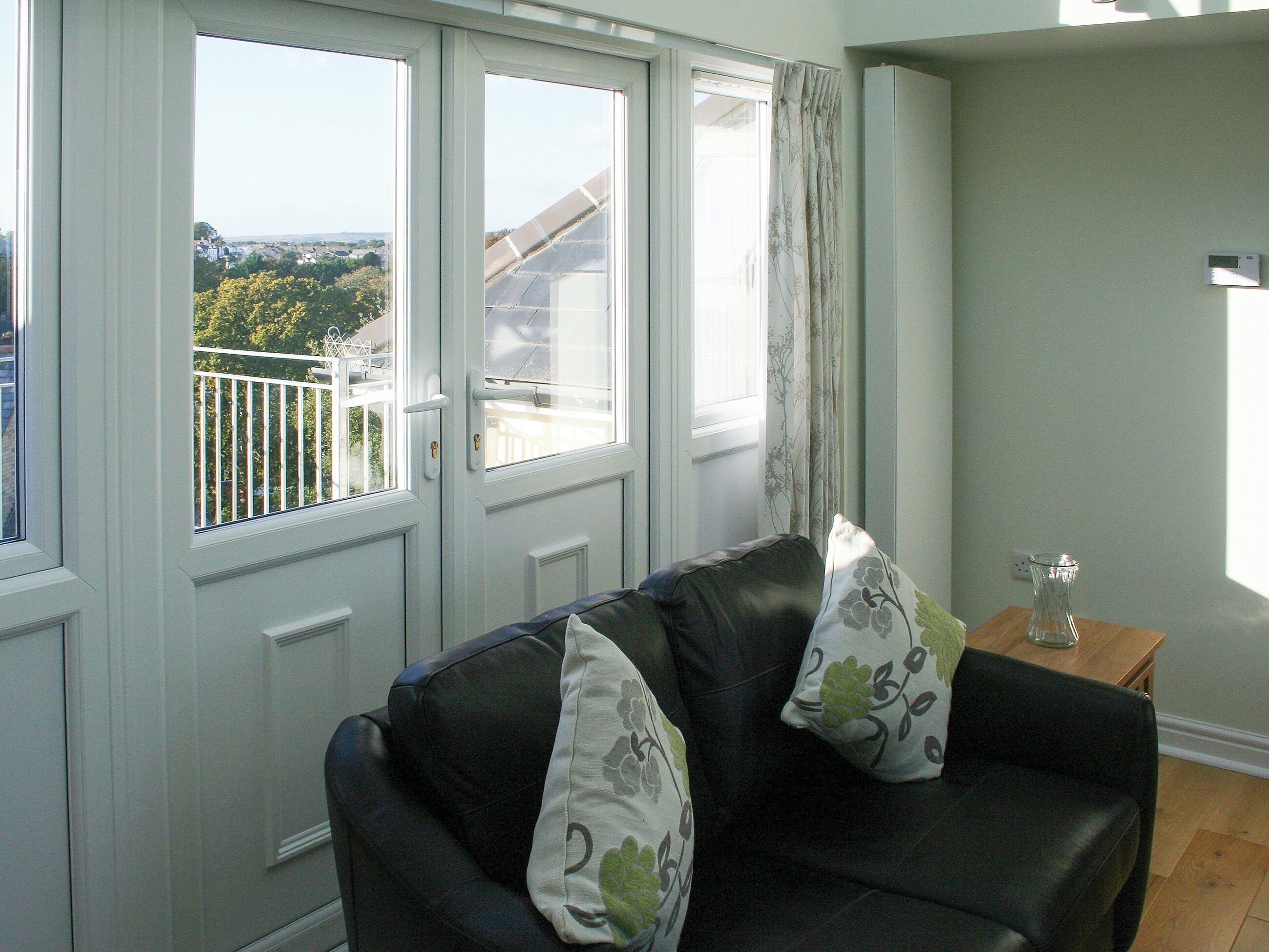 A living room with a black leather sofa, two cushions, a small wooden table with a glass vase, and a view of a balcony with a railing.