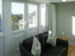 A living room with a black leather sofa, two cushions, a small wooden table with a glass vase, and a view of a balcony with a railing. - Highlands Apartment 4 - UKC3106 (Shanklin)