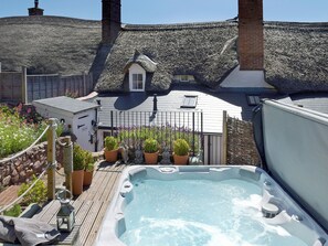 Un bain à remous sur une terrasse en bois, avec un mur en pierre et un bâtiment à toiture de chaume en arrière-plan.