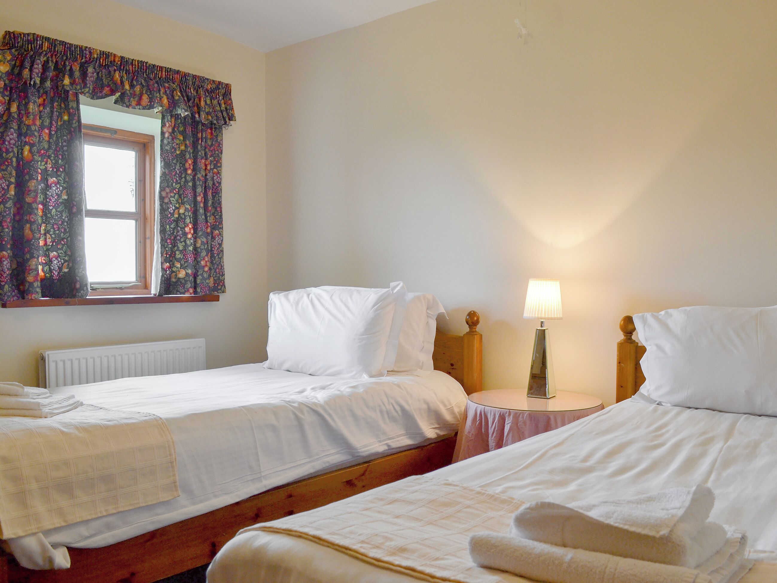A hotel room with two single beds, a window with floral curtains, a bedside table with a lamp, and a radiator.