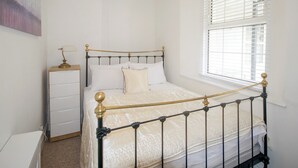 Ein ordentlich bezogenes Bett mit Metallgestell und Kopfteil, ein Nachttisch mit Lampe sowie ein Fenster mit Jalousien.
