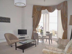 A modern living room with a flat-screen TV, a glass coffee table, a sofa, and a dining area with chairs. - This ground floor apartment is close to Torquay’s harbour, as well as Paignton. (Torquay)