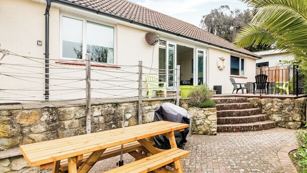A patio area with a wooden table and benches, a stone wall, and a house with a tiled roof and a door leading to a covered porch. - 3 bedroom accommodation in Ventnor (Ventnor)