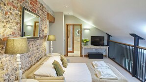 Une chambre d’hôtel avec un mur en pierre, un lit avec du linge de maison blanc, un canapé, un téléviseur posé sur un bureau et un balcon avec une balustrade.