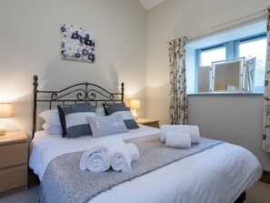 Ein ordentlich bezogenes Bett mit weißer und grauer Bettwäsche, zwei Nachttischlampen, ein Blumenbild an der Wand und ein Fenster mit Vorhängen.
