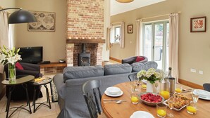 A living room with a brick fireplace, grey sofas, a dining table set with breakfast, and a TV in the corner. - The Milking Parlour - UKC3681 (Tattersett, near Fakenham)