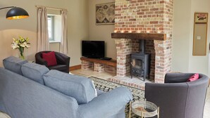 A living room with a brick fireplace, a sofa, a television, and a lamp. - The Milking Parlour - UKC3681 (Tattersett, near Fakenham)