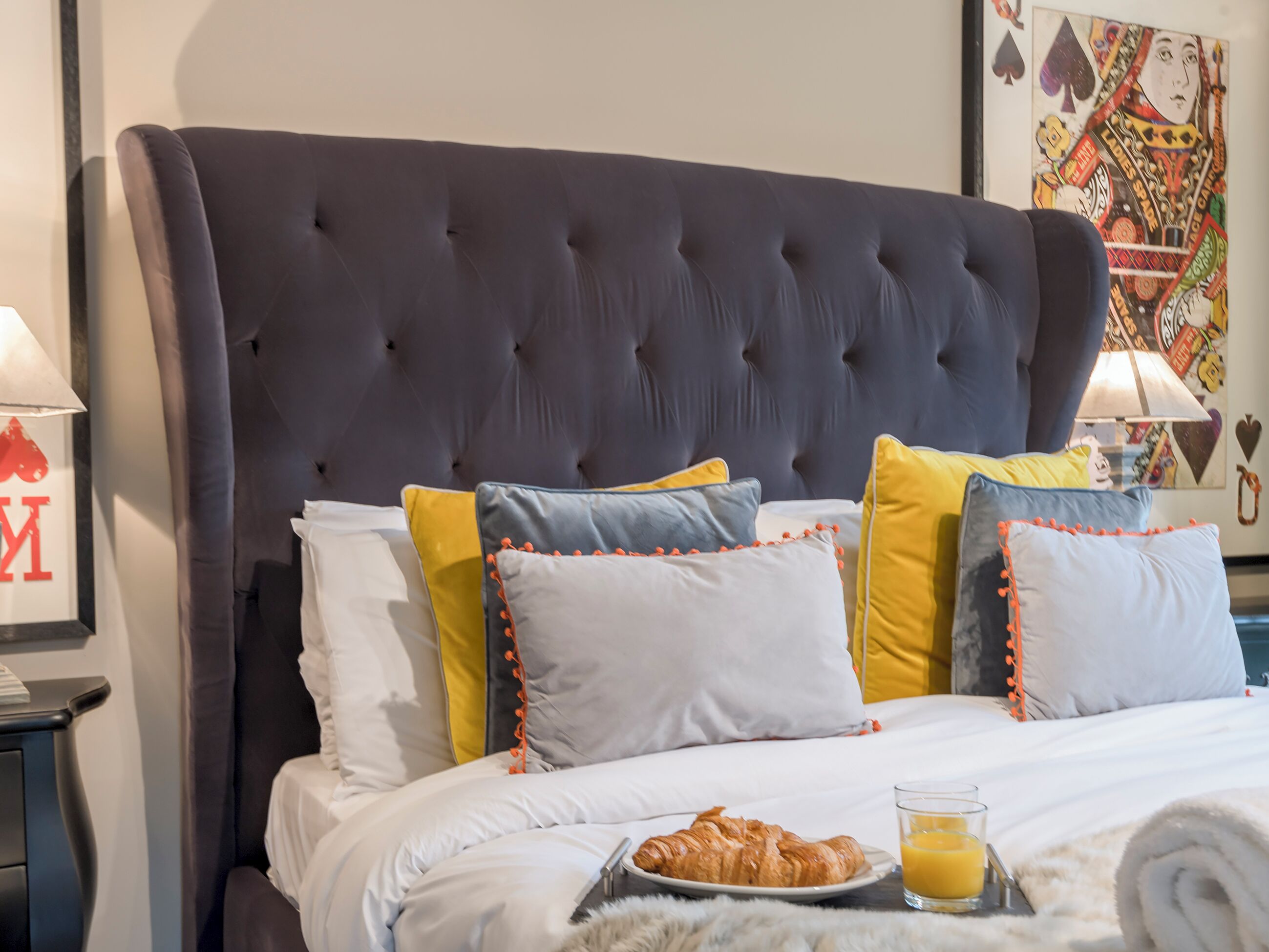 A neatly made bed with a dark headboard, multiple pillows in various colors, a plate with croissants, and a glass of orange juice on the bedside table.