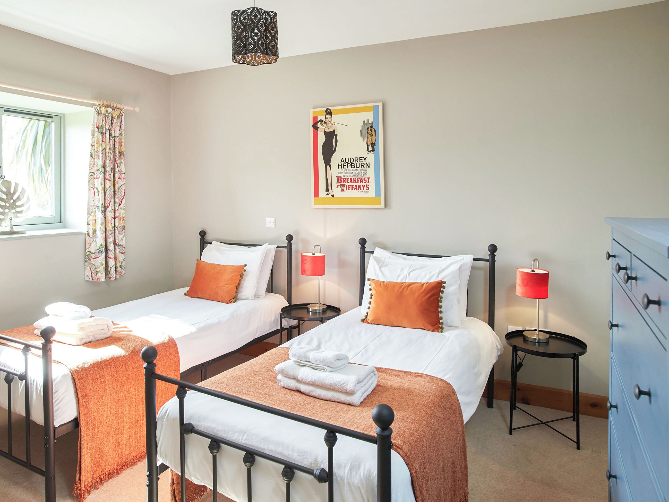 Une chambre double avec deux lits, chacun agrémenté de draps blancs et d’oreillers orange, une petite table avec une lampe rouge et une commode.