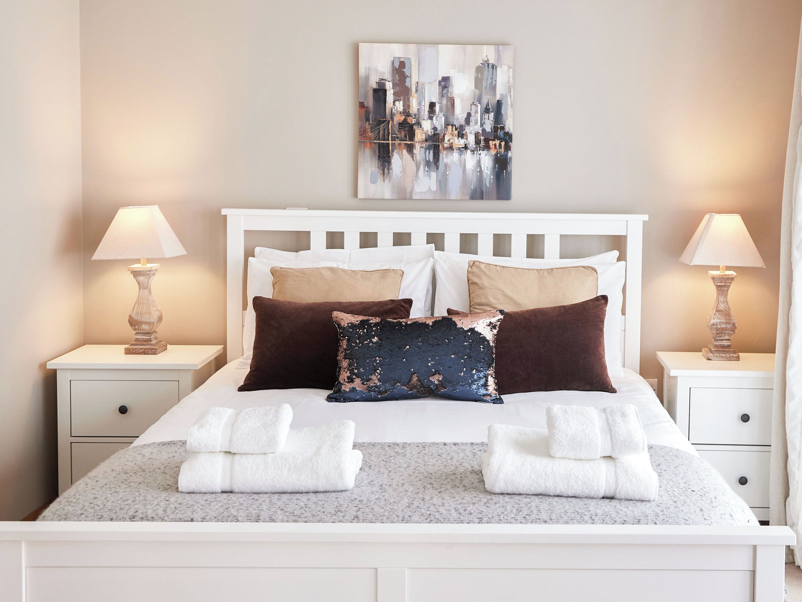Una cama bien hecha con sábanas blancas y dos almohadas, un cuadro de la ciudad sobre ella, y dos lámparas de noche en mesitas blancas.