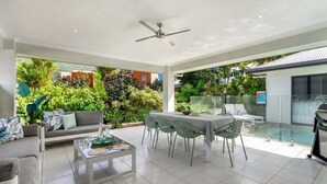 Ett modernt vardagsrum med en soffa, ett soffbord, ett matbord och stolar, en takfläkt och utsikt över en swimmingpool och grönområden.