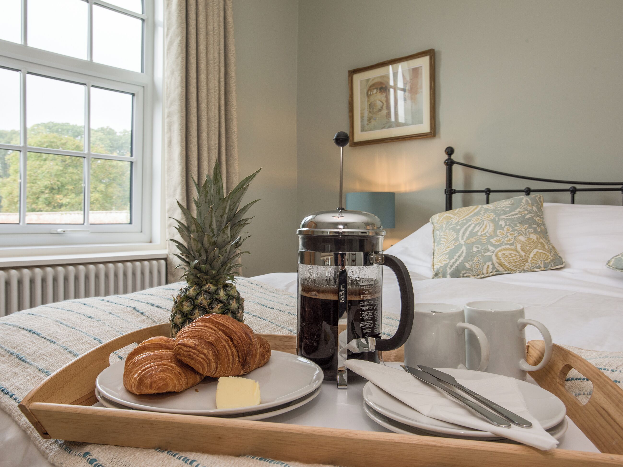 Un plateau de petit-déjeuner avec des croissants, du beurre, une cafetière et une tasse, posé sur un lit d’où l’on aperçoit les arbres à travers une fenêtre.