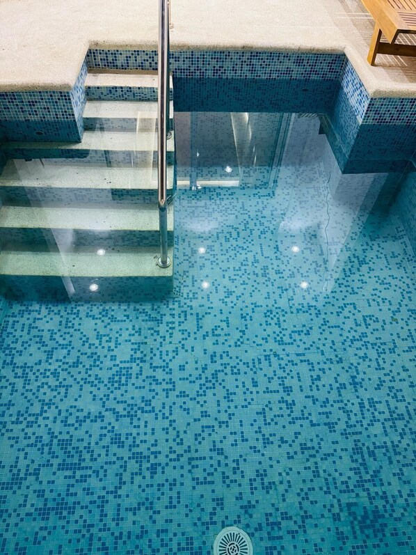Une piscine aux carreaux de mosaïque bleus et dotée d’une barrière en verre avec des rampes métalliques.