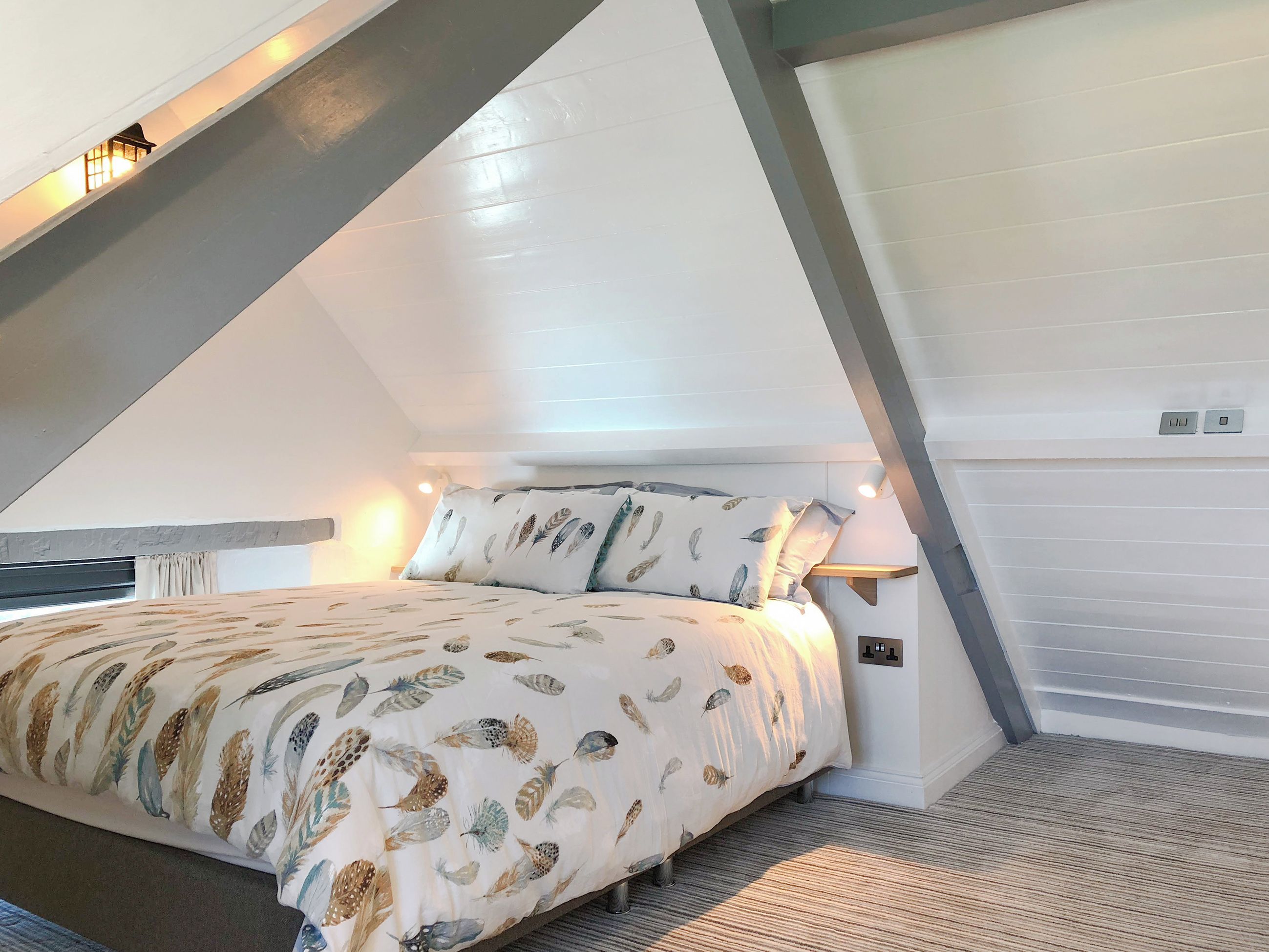 A bedroom with a sloped ceiling, a bed with feather-patterned bedding, and a small window with curtains.