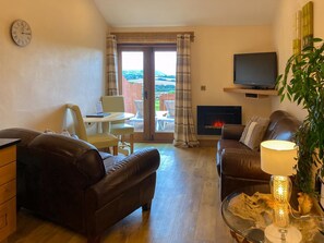 A living room with a fireplace, leather sofas, a TV, and a dining area with a laptop. - 1 bedroom accommodation in Marhamchurch, near Bude (Marhamchurch, near Bude)