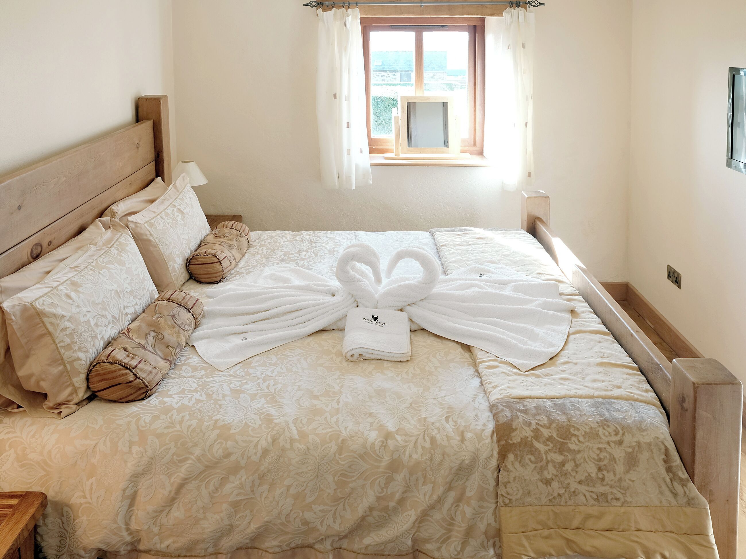 A neatly made bed with a towel arranged in a heart shape, a wooden headboard, and a window with curtains.