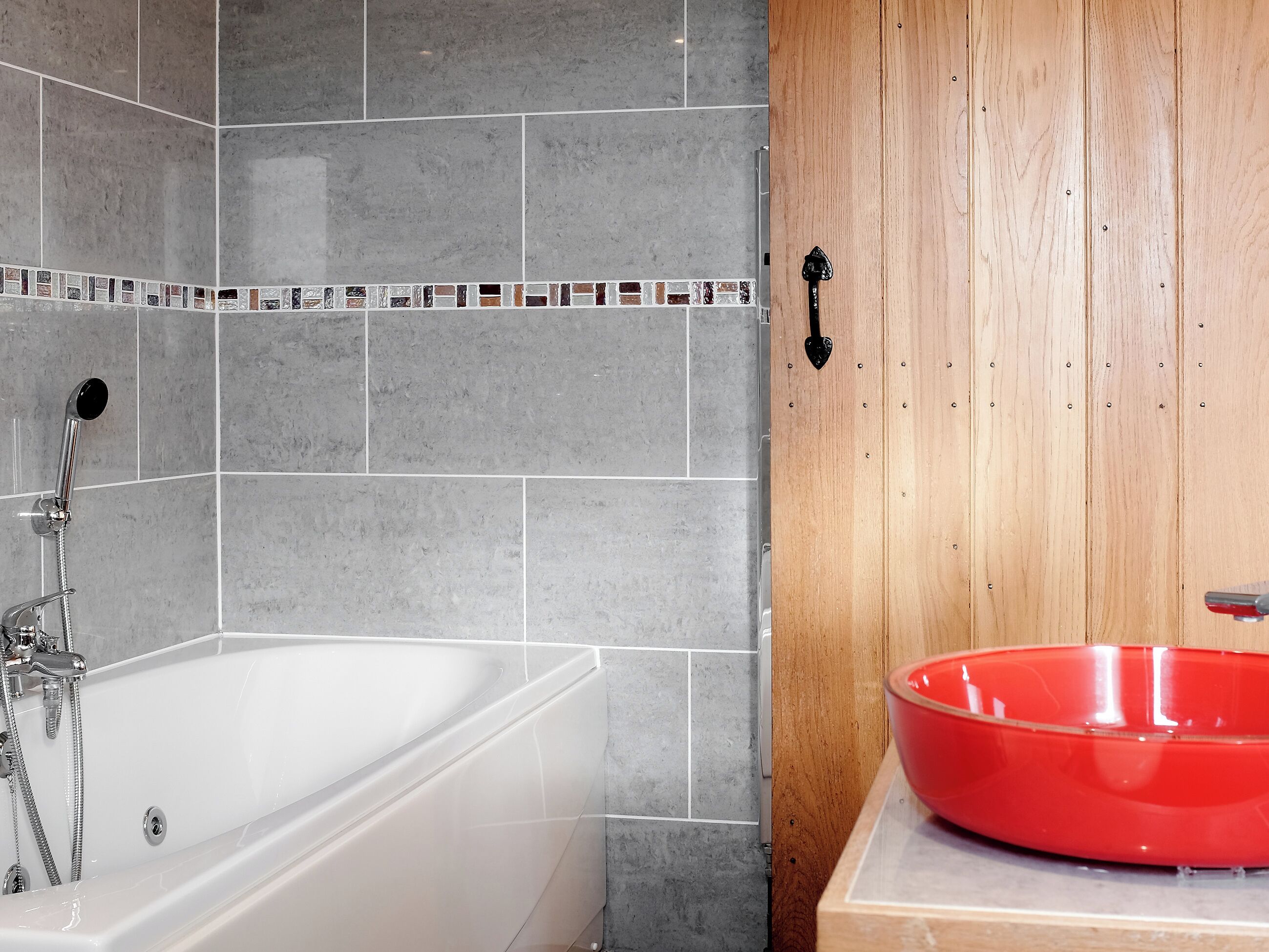 Une salle de bain avec une baignoire blanche, des murs recouverts de carreaux gris, une porte en bois et un lavabo rouge.