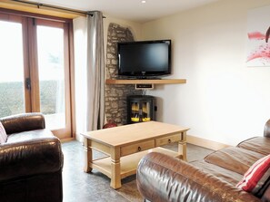 A living room with a television, a stone wall, a fireplace, leather sofas, and a wooden table. - 1 bedroom accommodation in Marhamchurch, near Bude (Marhamchurch, near Bude)