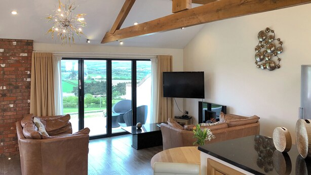 A spacious living area with a fireplace, leather sofas, a TV, and a large window with a view of the countryside. - 1 bedroom accommodation in Marhamchurch, near Bude (Marhamchurch, near Bude)
