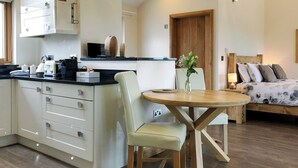 A modern kitchen with white cabinets, a dining area with a round table and chairs, and a bedroom with a bed and pillows. - 1 bedroom accommodation in Marhamchurch, near Bude (Marhamchurch, near Bude)