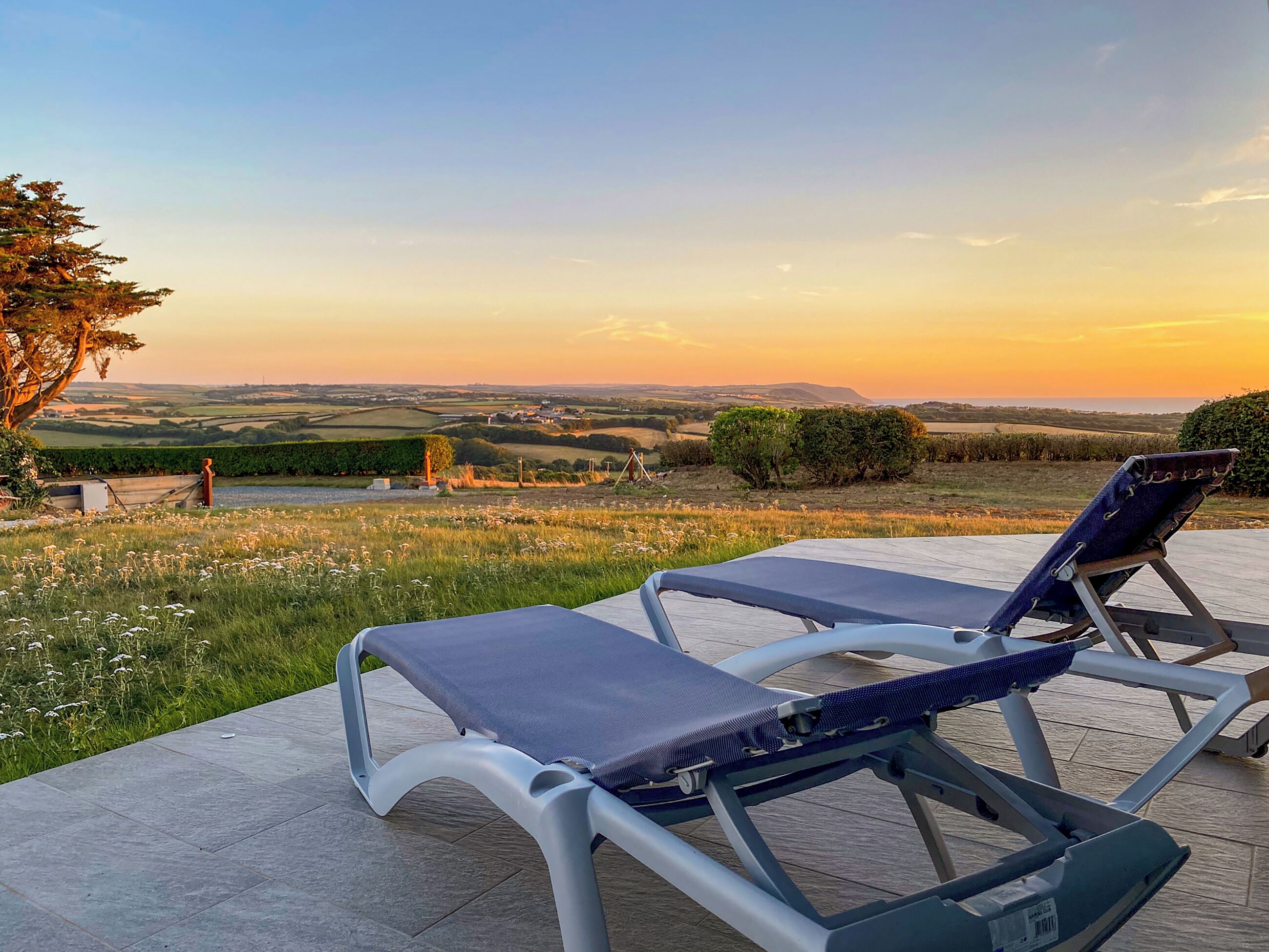 Two lounge chairs on a patio with a view of rolling hills and a sunset.