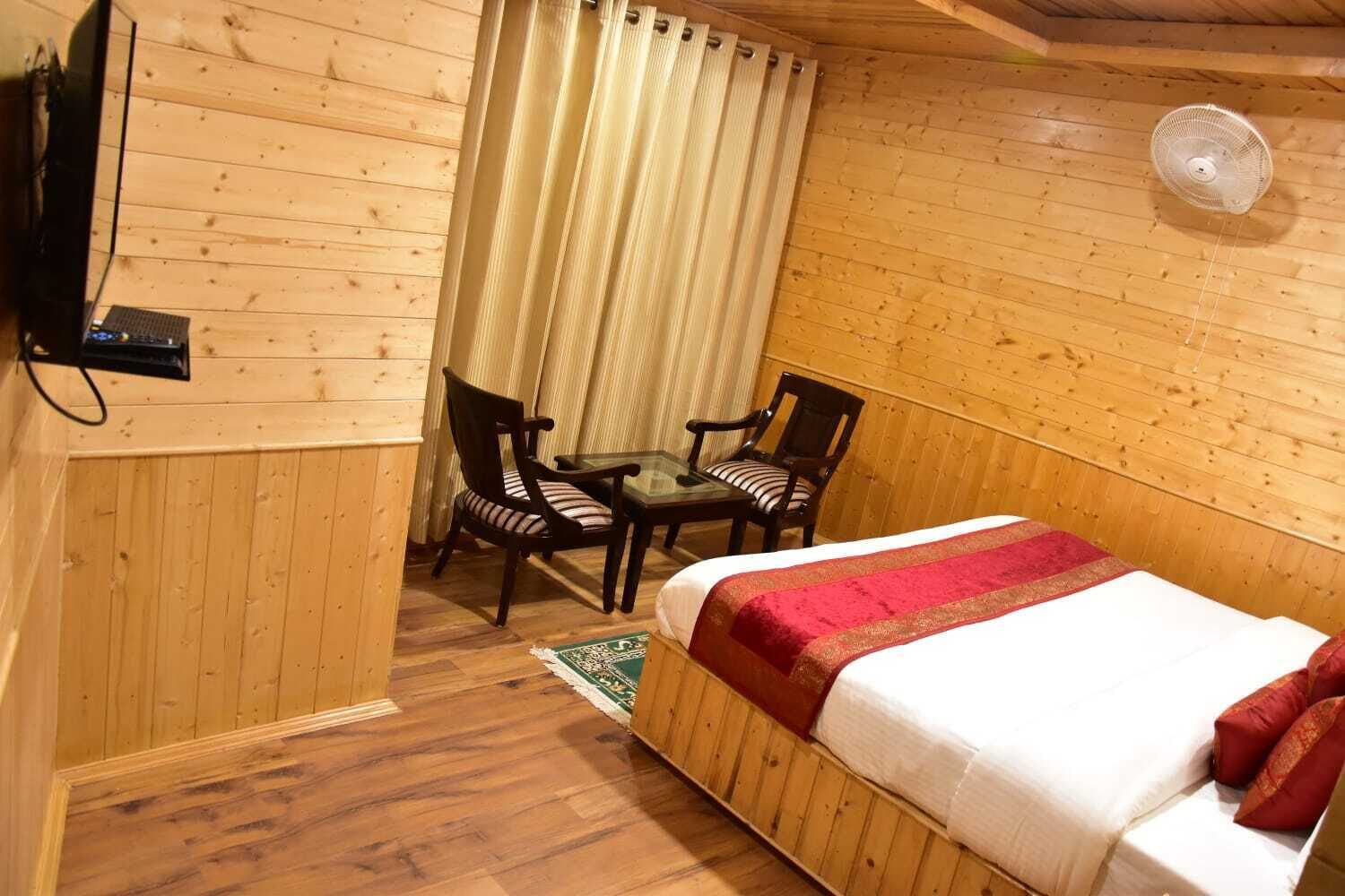 A wooden cabin room with a bed, two chairs, a small table, and a fan.